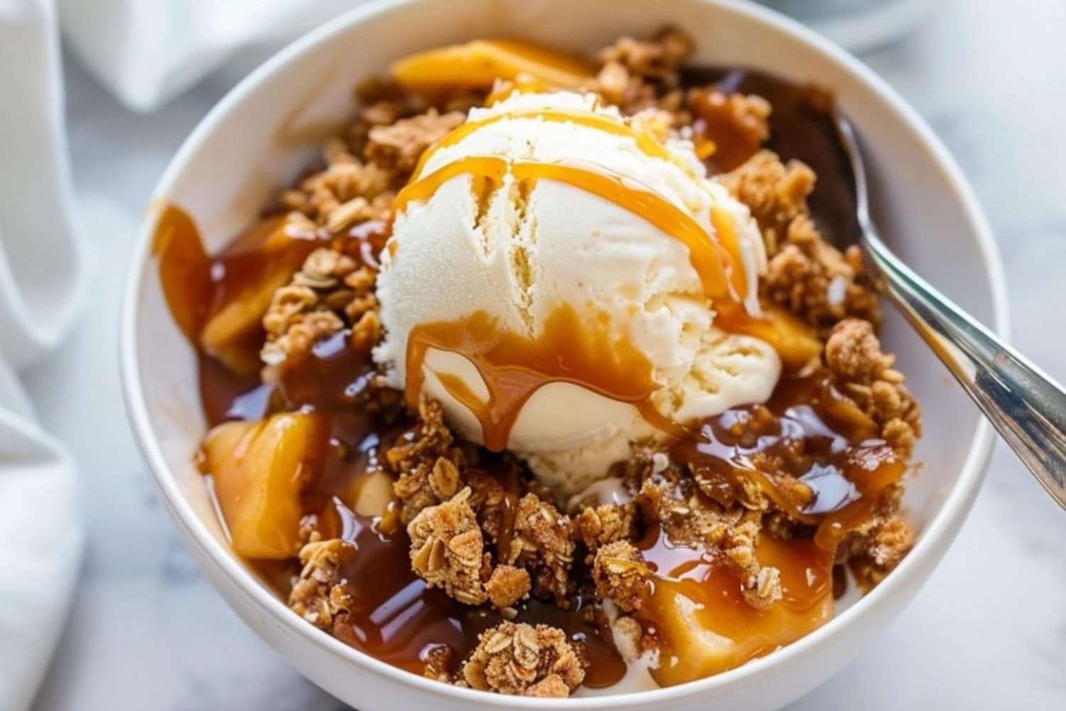 Caramel apple crisp in a white bowl with scoop of vanilla icing and a piece of fork on the side.