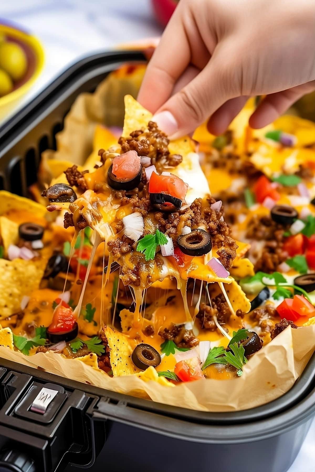 Hand lifting a piece of cheesy nachos from air fryer.