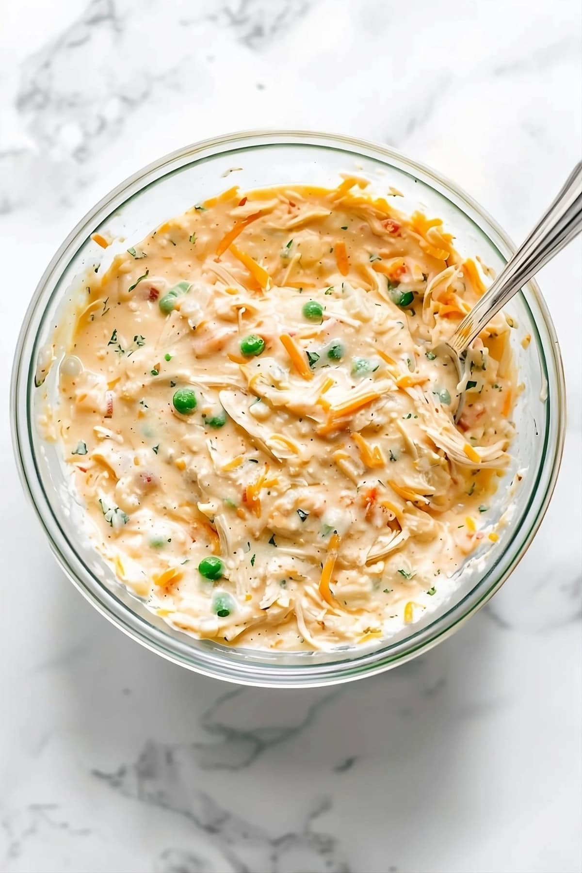 Bowl with a mix of shredded chicken, cream of chicken soup, sour cream, milk, diced onion, peas and carrots, garlic powder, parsley, salt, pepper, and cheddar cheese.
