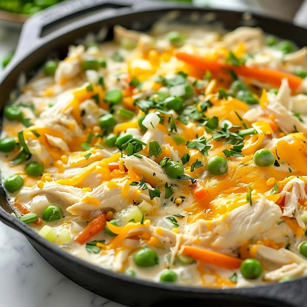 Chicken and biscuit casserole filling in a cast iron skillet with shredded chicken, frozen peas, and cheddar cheese, carrots and chopped parsley.