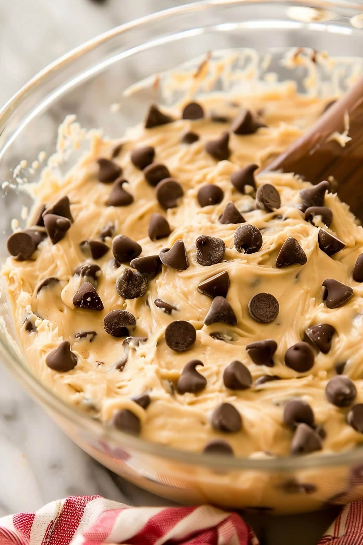 A glass bowl of chocolate chip cookie dough with a wooden spoon