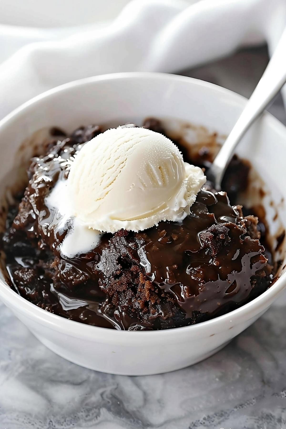 Chocolate cobbler in a white bowl garnished with scoop of vanilla ice cream with spoon on the side.