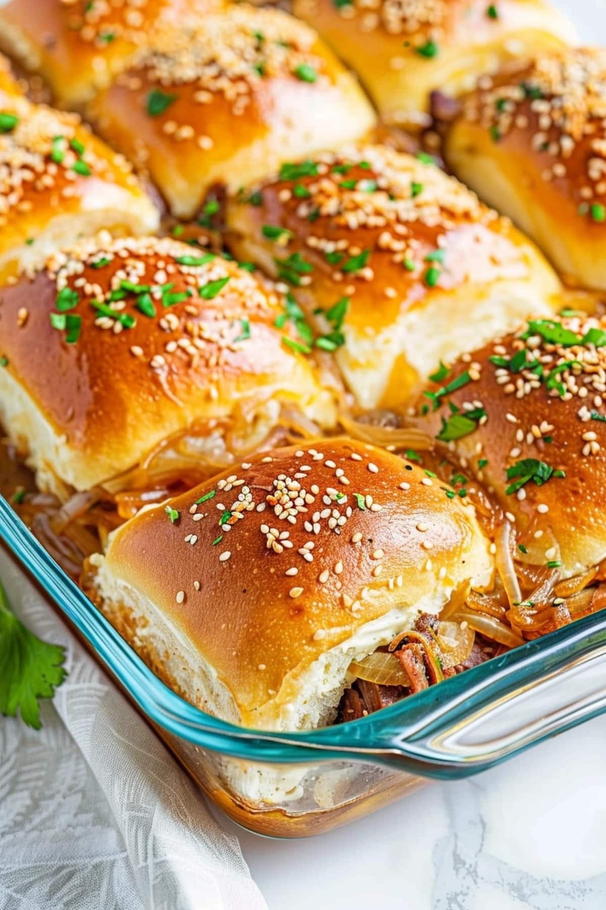 French dip sliders in glass baking dish topped with sesame seeds and chopped parsley.