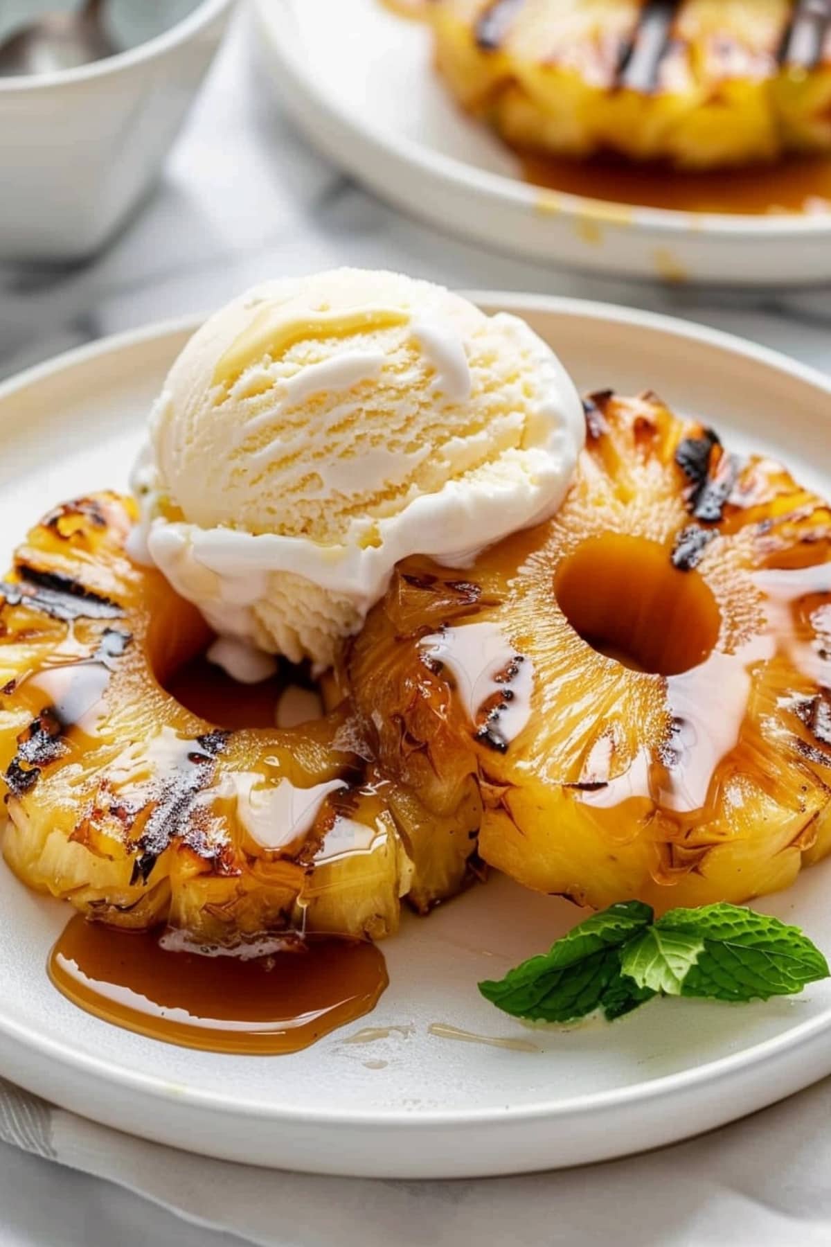 Two slices of grilled pineapple rings with scoop of vanilla ice cream on top and a mint sprig on the side