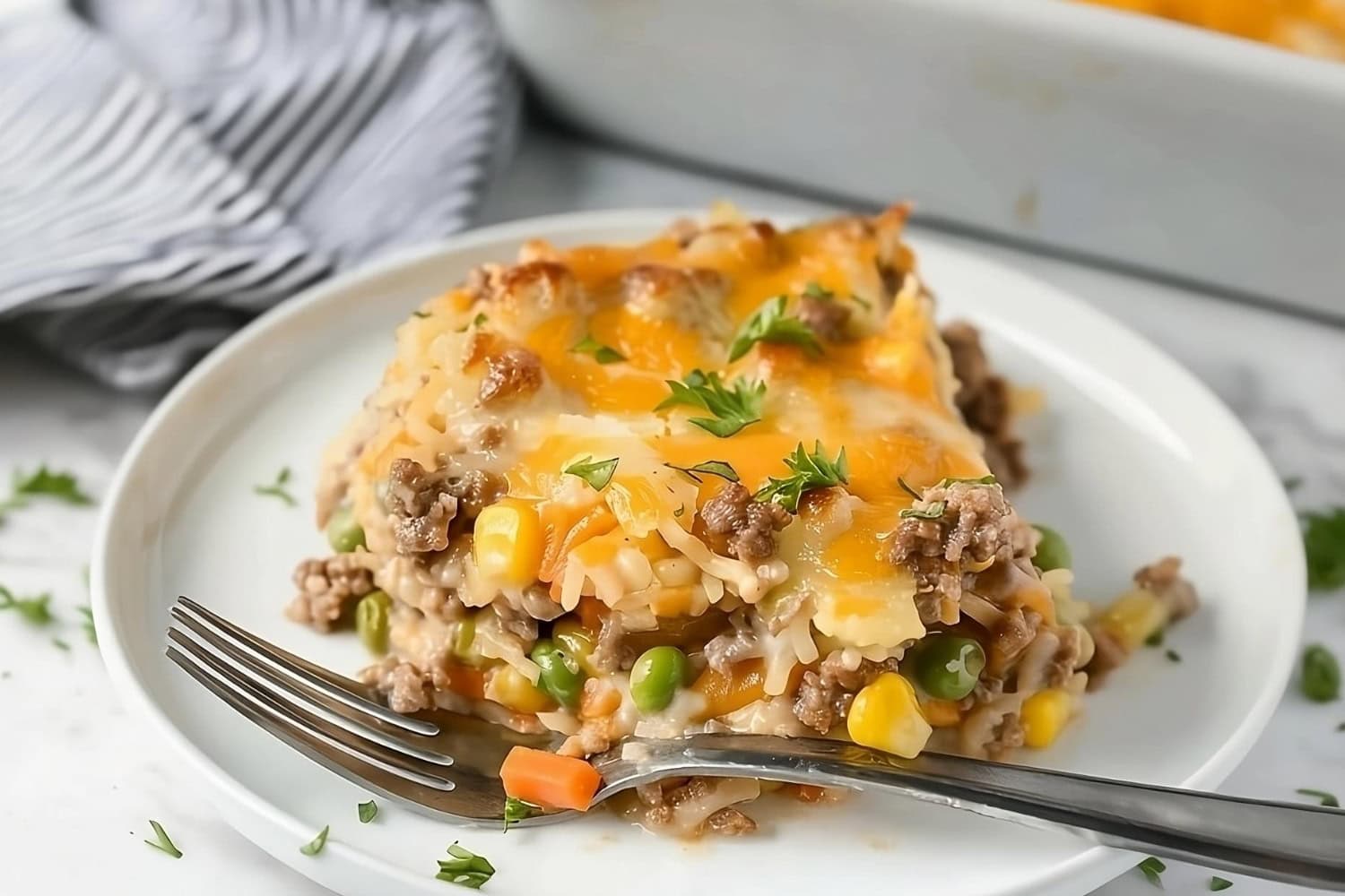 Slice of hamburger hashbrown casserole in plate with carrots, peas, and corn.