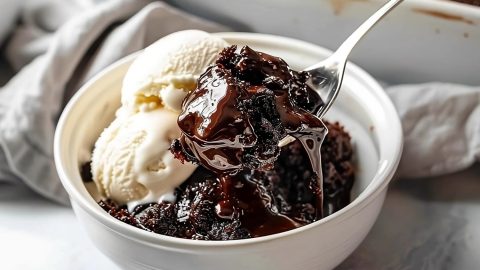 Spoon scooping a serving of hot fudge cake with ice cream served on a white bowl.