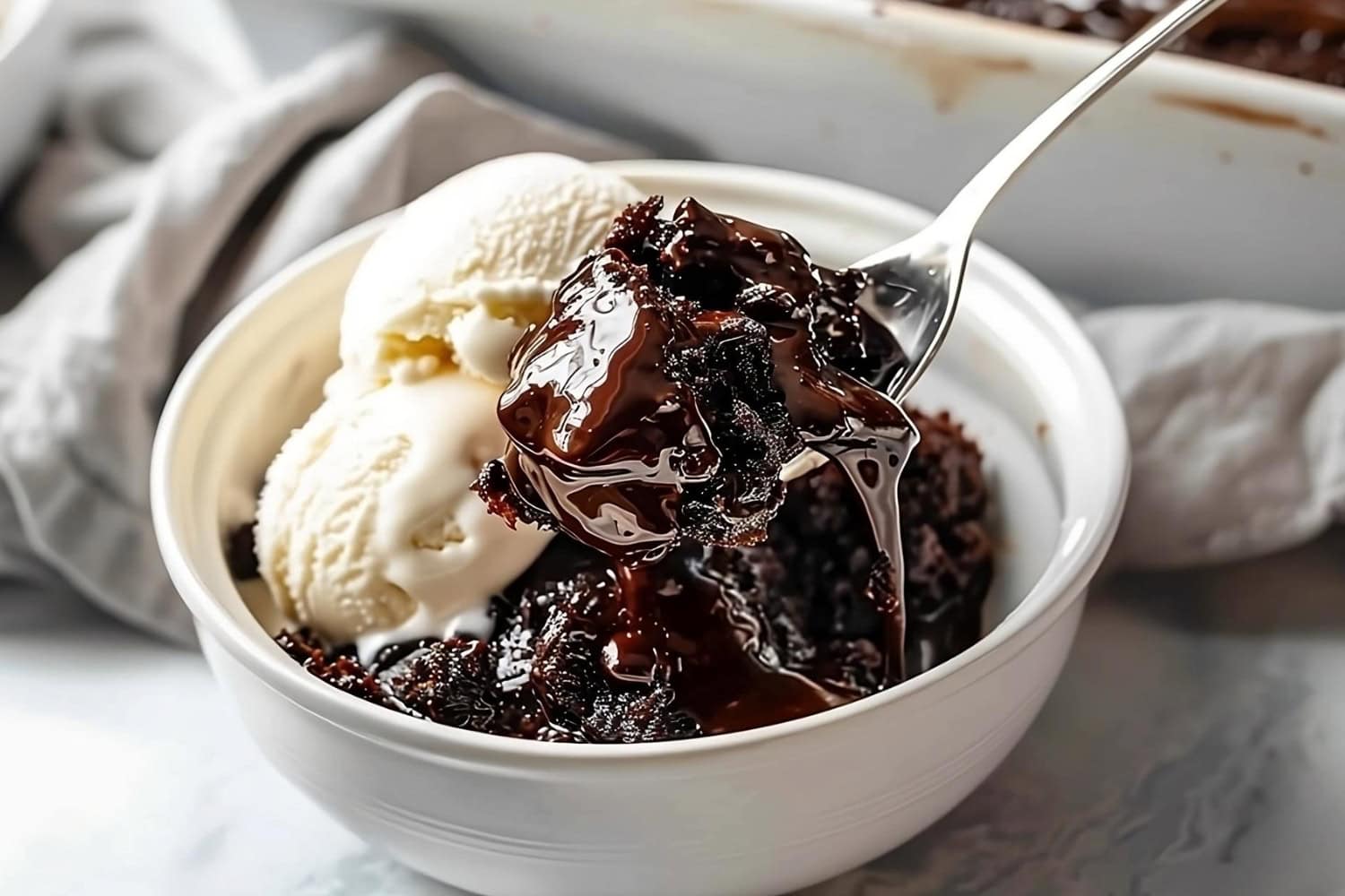 Spoon scooping a serving of hot fudge cake with ice cream served on a white bowl.