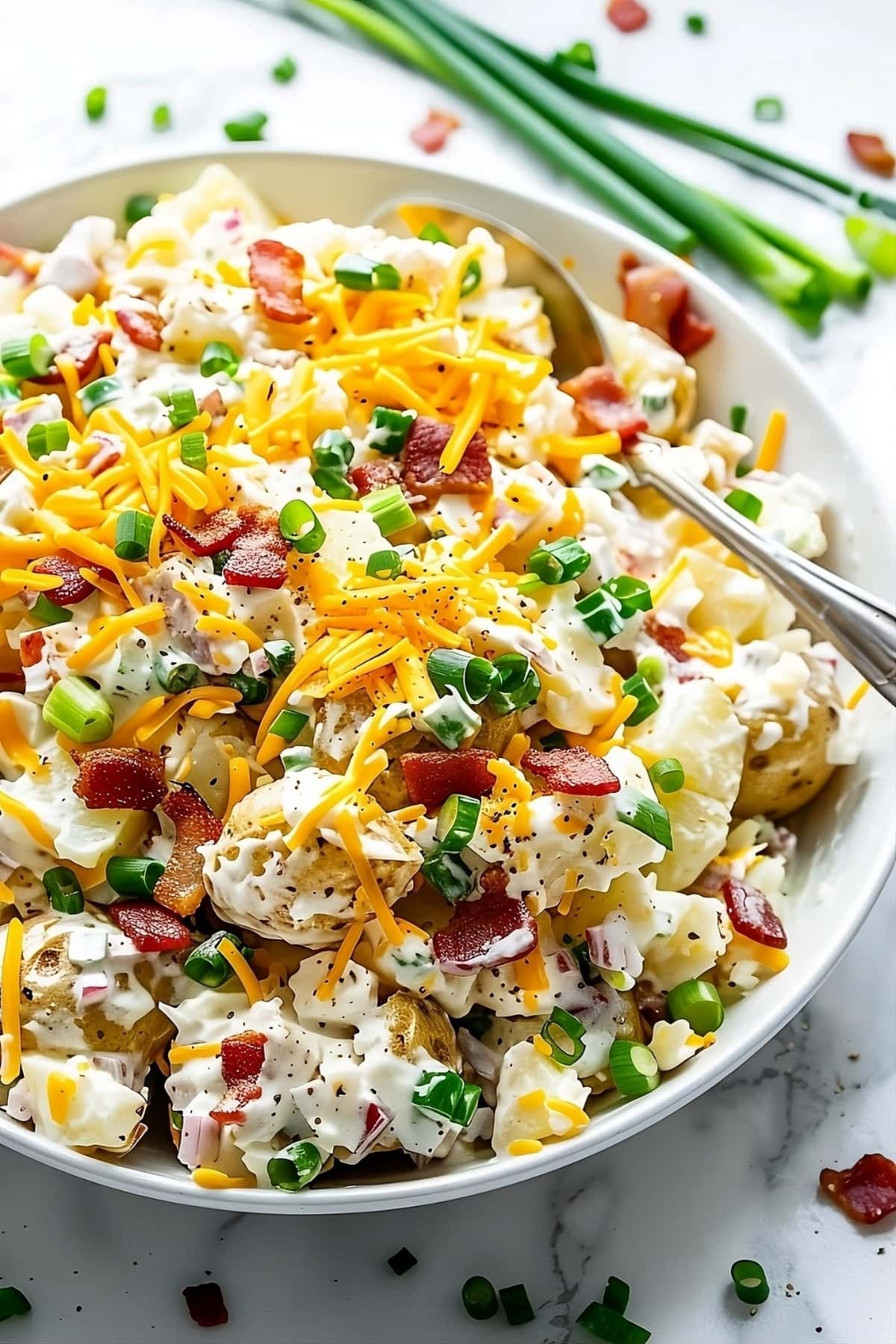 Loaded baked potato salad topped with lots of shredded cheese, bacon and green onions in a white bowl with spoon.