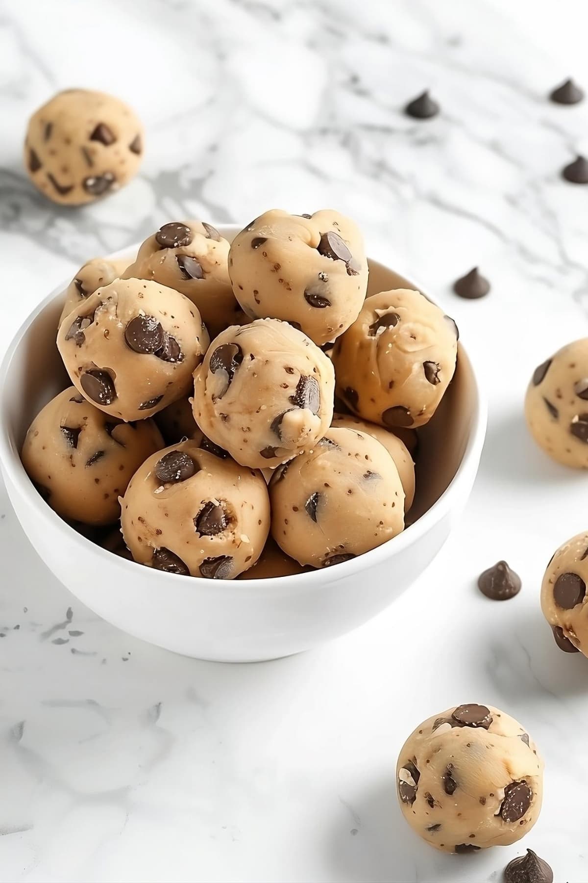 No bake cookie dough balls in a white bowl on a white marble surface.