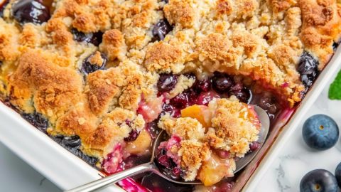 Spoon scooping peach and blueberry cobbler from baking dish.