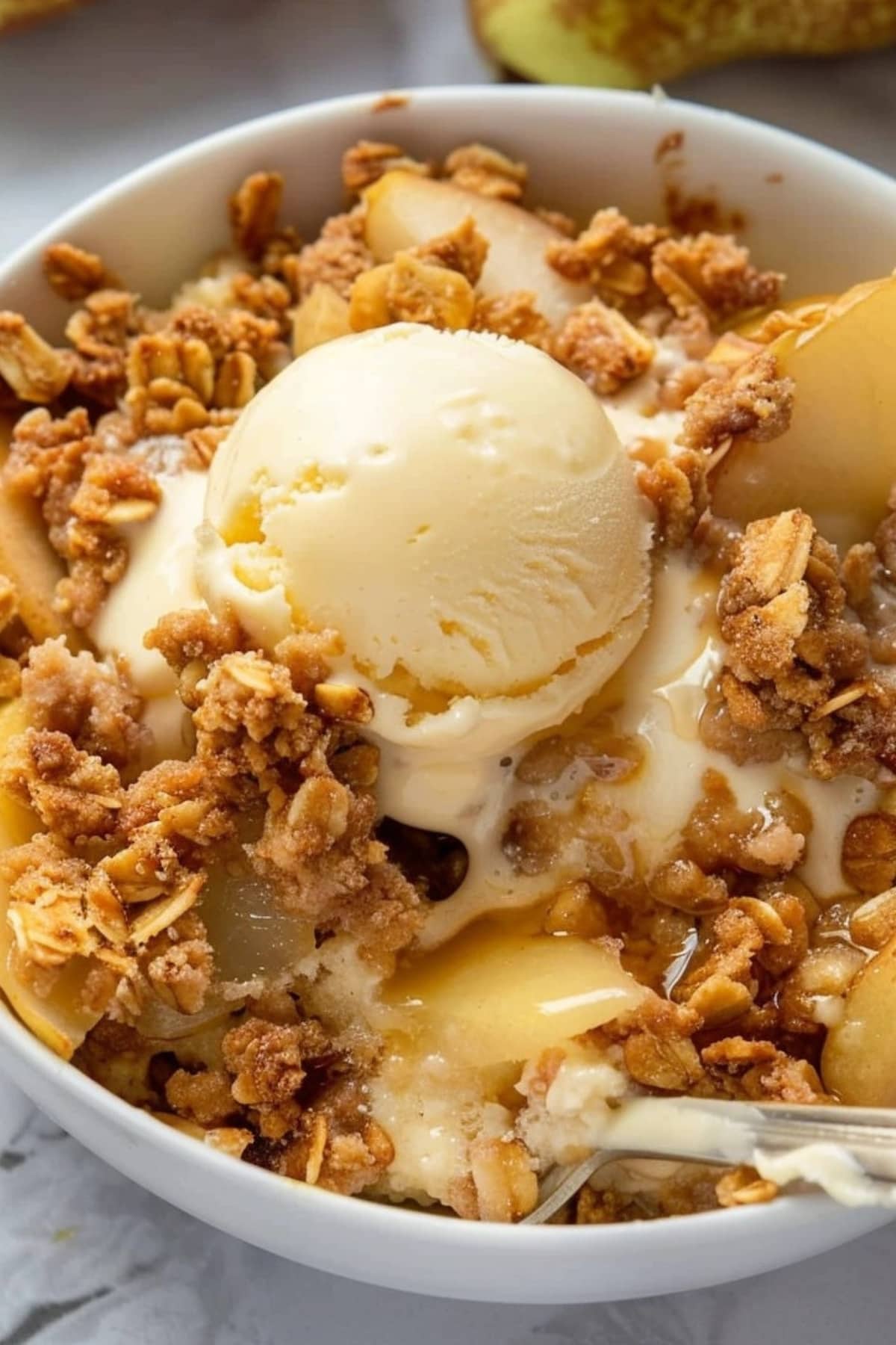Pear crisp served in a white bowl topped with scoop of vanilla ice cream.