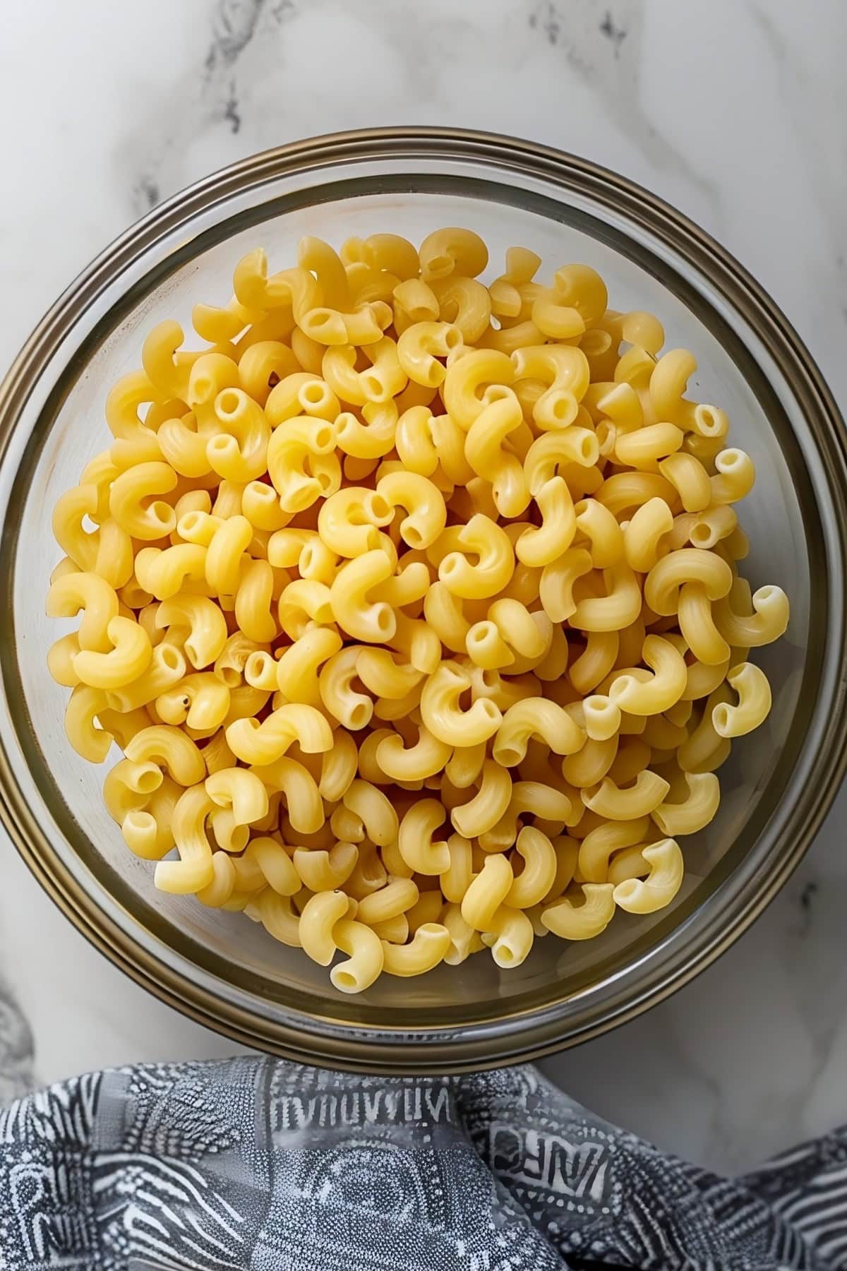 A glass bowl of uncooked elbow macaroni pasta on a white marble table