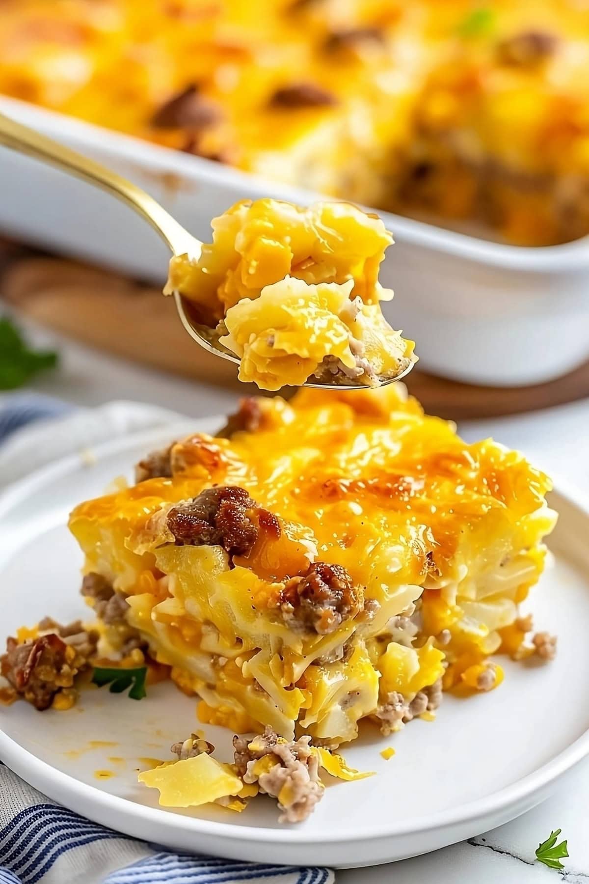 Spoonful of sausage hashbrown breakfast casserole on a plate with a white baking dish in the background.