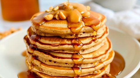 Stack of peanut butter pancake drizzled with maple syrup garnished with peanut butter and peanuts.