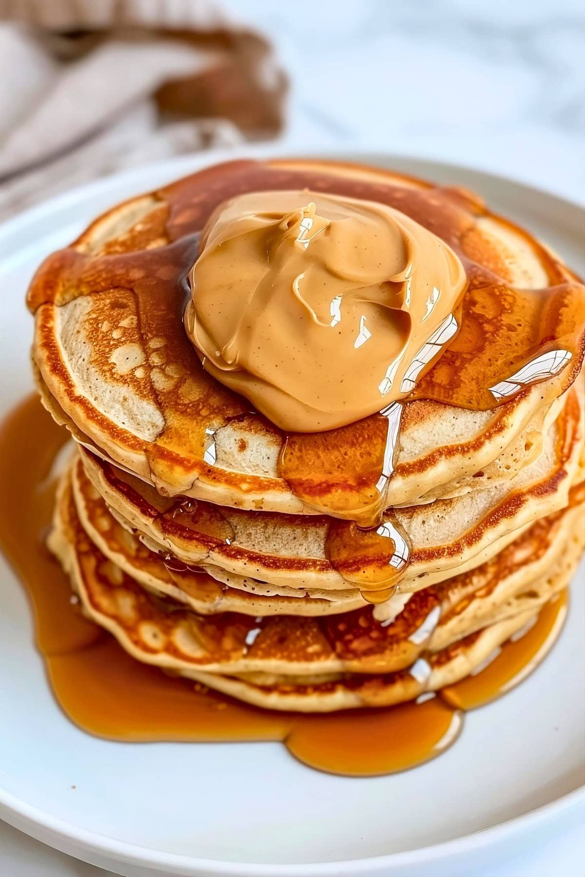 Stack of peanut butter pancakes dripping with maple syrup in a plate with peanut butter spread on top.