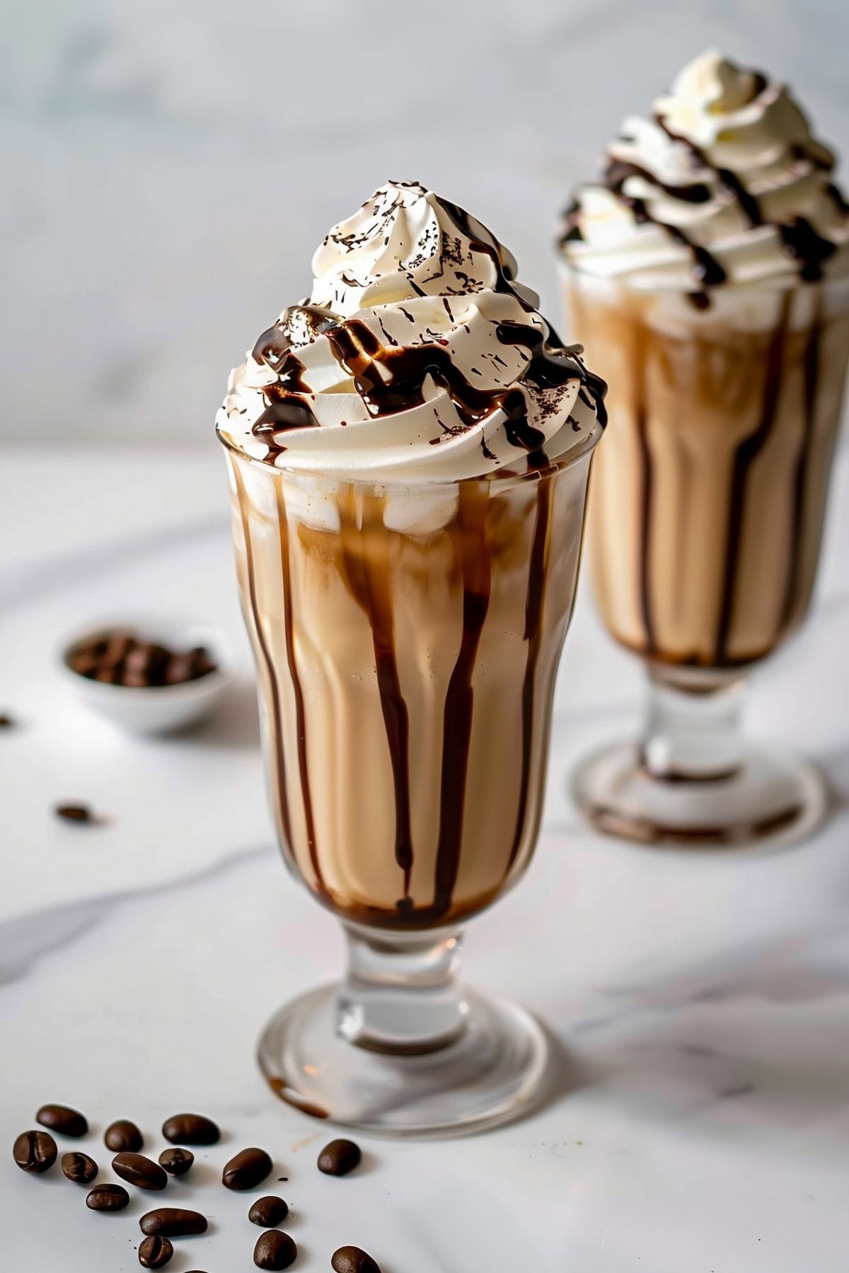 Two glasses of coffee milkshake topped with whipped cream, drizzled with chocolate sauce