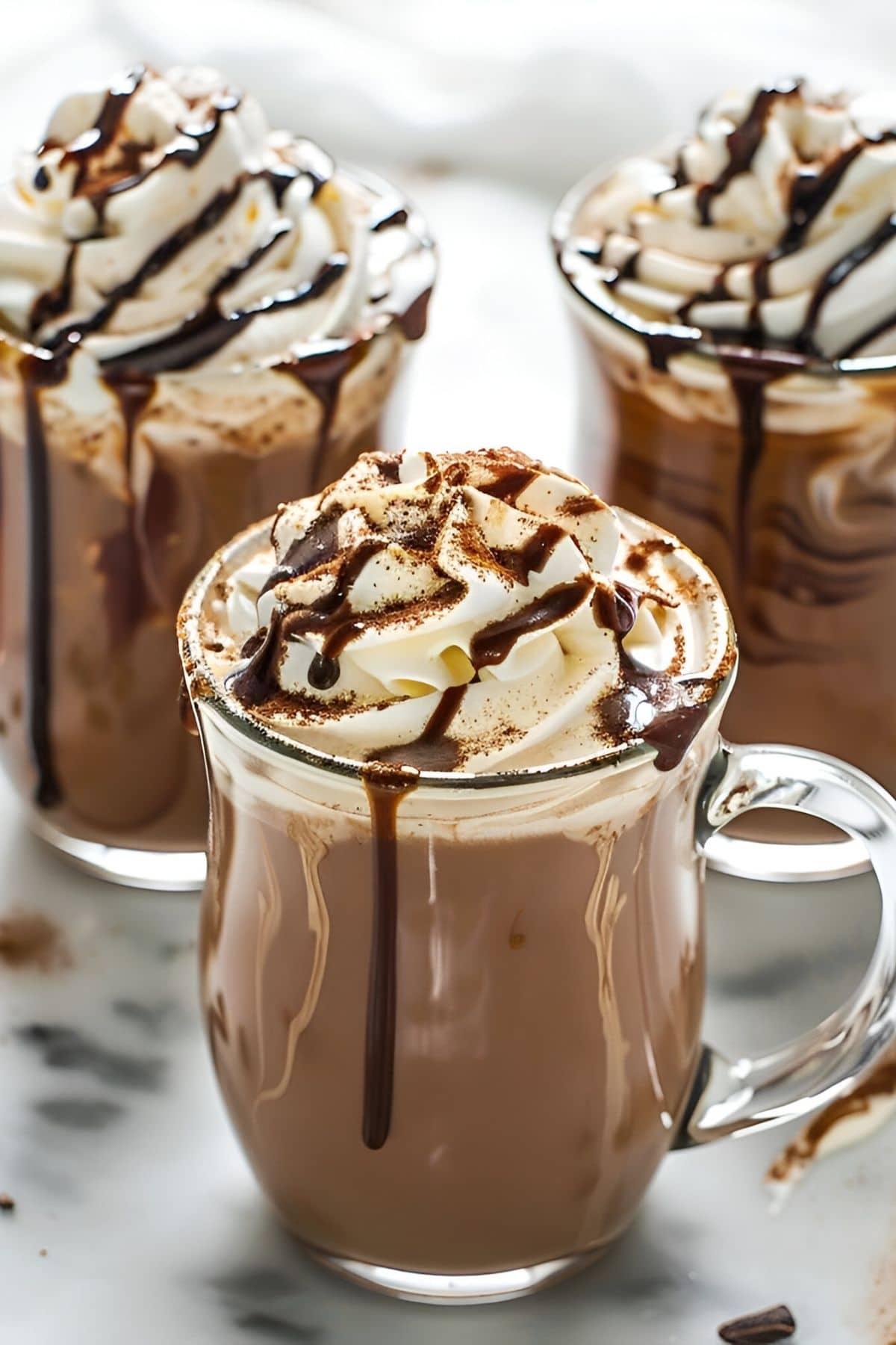 Three Glass Mugs of Bailey's Hot Cocoa with Whipped Cream, Chocolate Drizzle, and Cocoa Powder on a White Counter