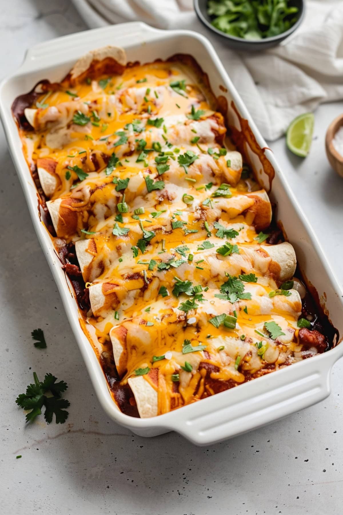 Baking dish with beef enchiladas with a bowl of chopped parsley and a slice of lime on a marble table.