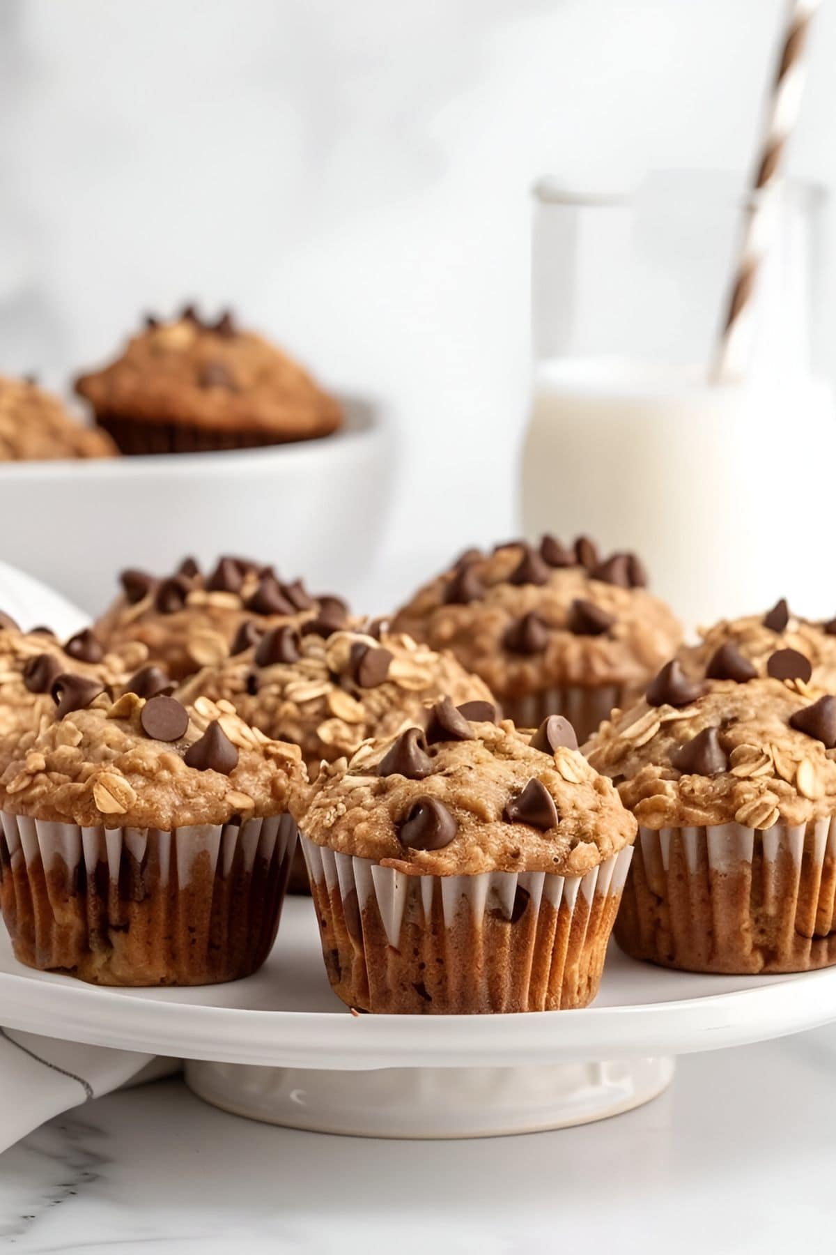 Chocolate Chip Oatmeal Muffins on a Cake Plate with More Muffins in a Bowl in the Background and a Glass of Milk with a Straw