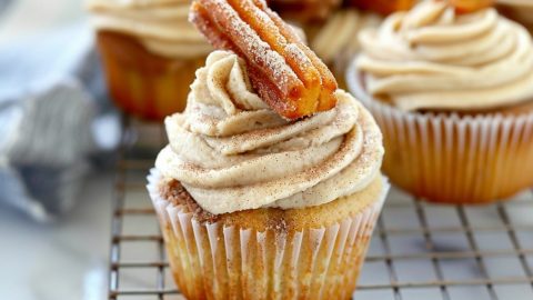 Cupcakes with cinnamon frosting garnished with a slice of churro arranged in a while cooling rack.