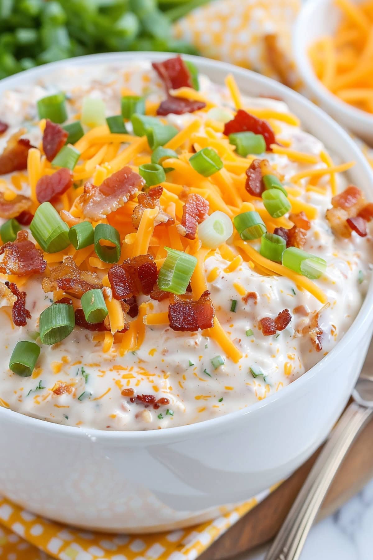 Crack dip in a white bowl topped with chopped fried bacon, green onions and cheddar cheese.