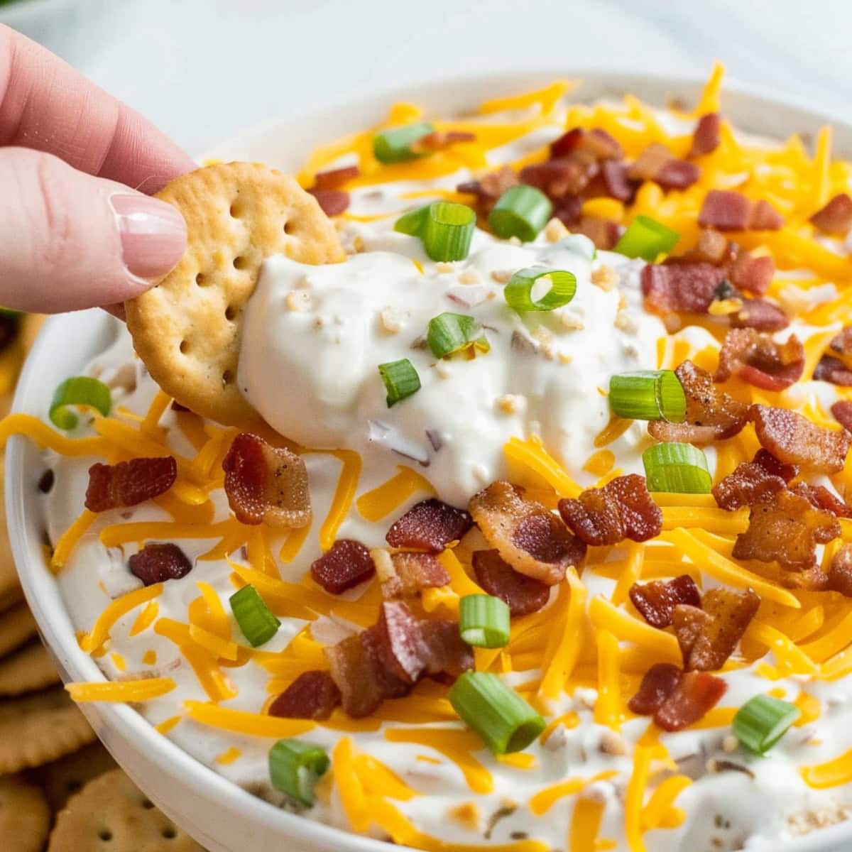 Crackers dipped in a creamy dip made with mixture of sour cream and ranch dressing with crumbled crispy bacon bits, shredded cheddar cheese, sliced green onions on a white bowl