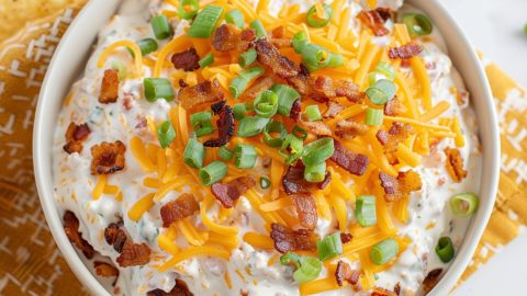 A bowl of crack dip garnished with shredded cheddar cheese, bacon bits and green onions.