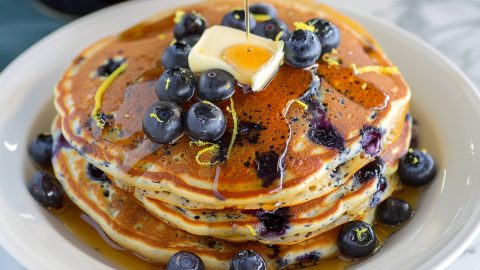 Comforting lemon blueberry pancakes, stacked on a plate and served with a generous pour of warm maple syrup.