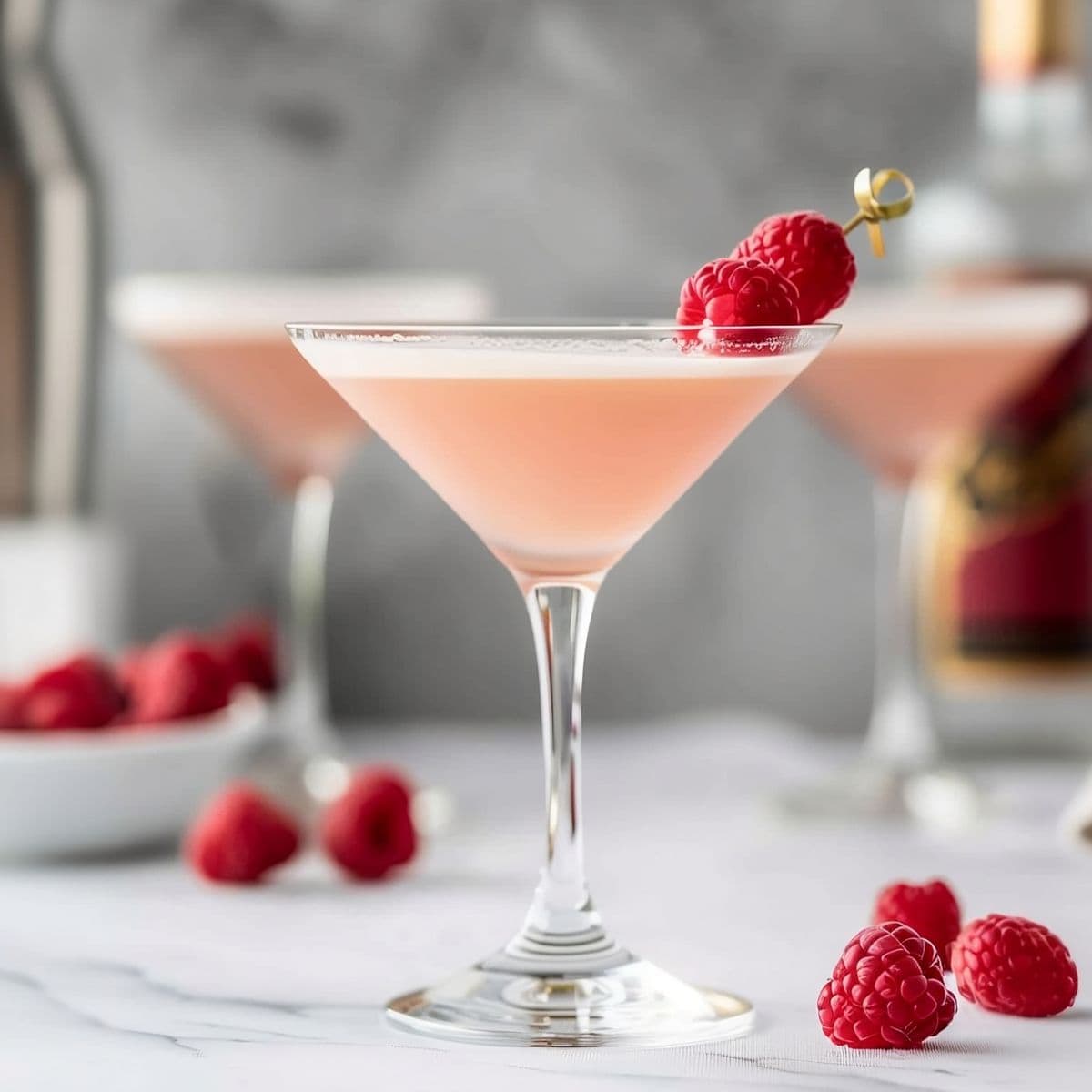 Three French Martinis with Raspberries on a White Marble Table with a Bottle of Chambord, a Cocktail Shaker, and More Raspberries in the Background