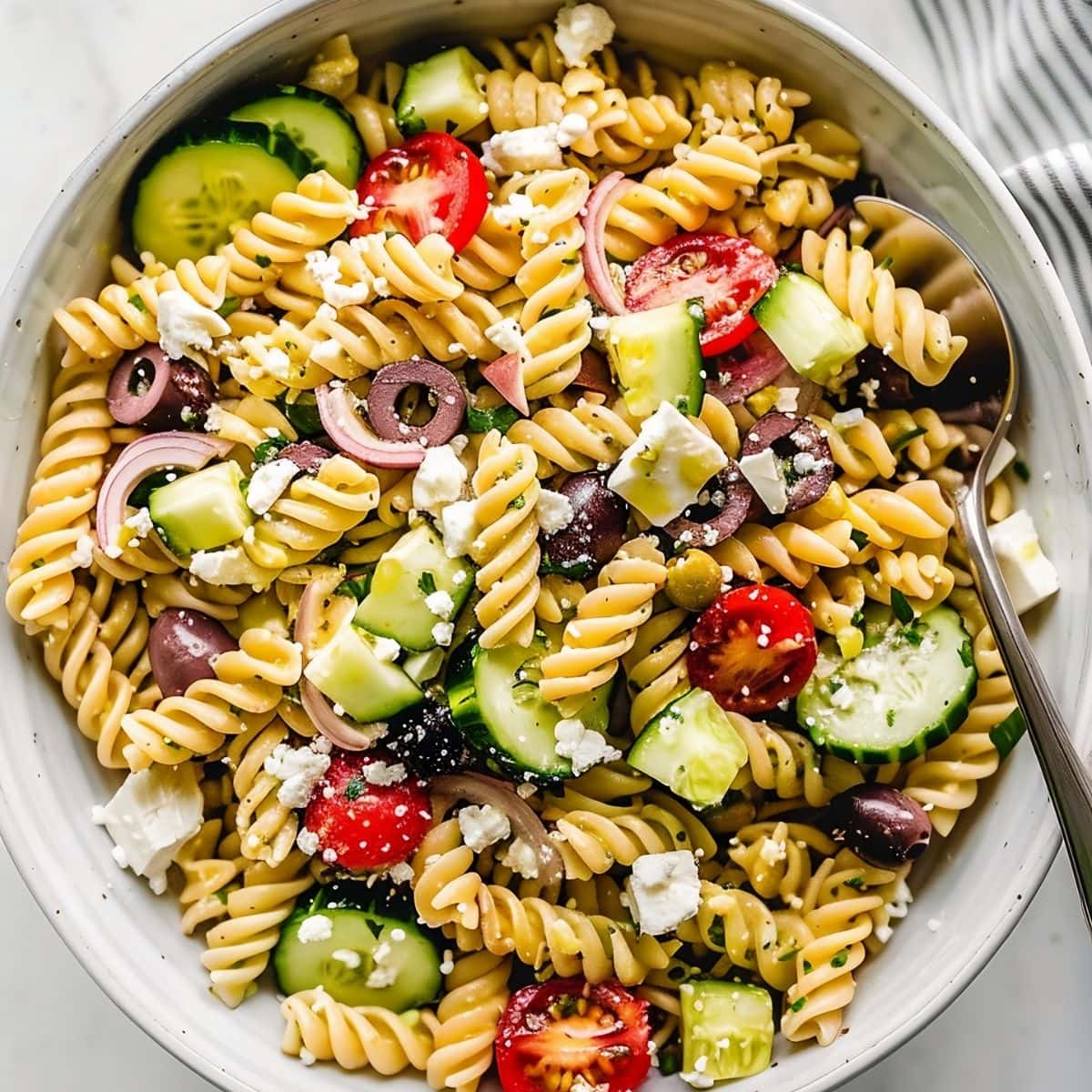Top View of Bowl of Greek Pasta Salad with Olives, Red Onions, Cucumber Slices, Tomatoes, Feta, and Herbs with a Spoon 