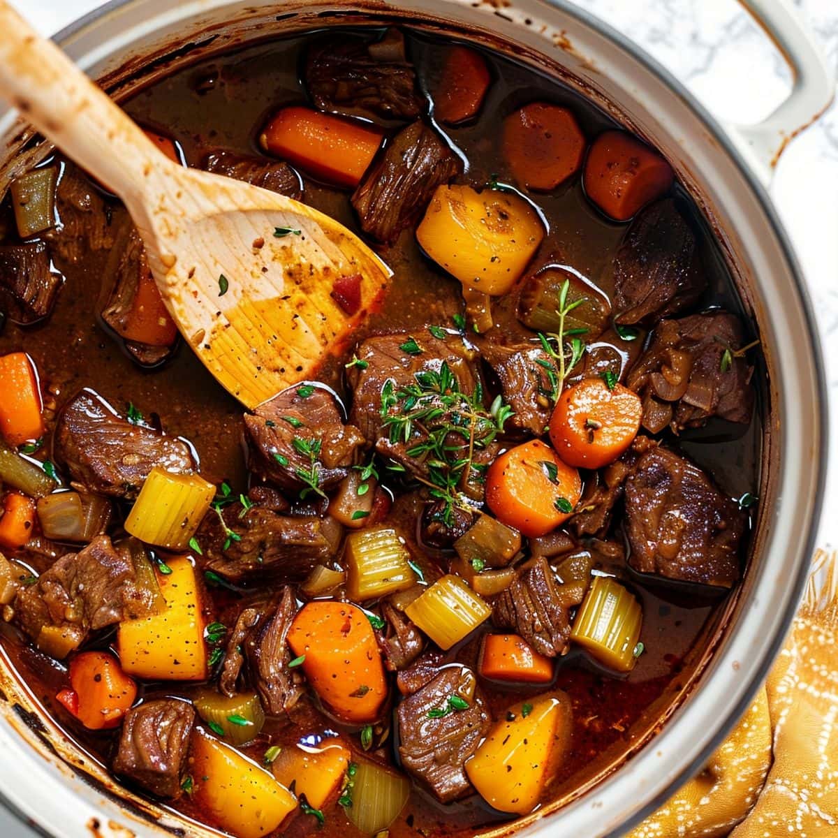 Top View of Guinness Beef Stew with Meaty Beef, Carrots, Celery, and Herbs in a White Dutch Oven with a Wooden Spoon on a White Marble Table with a Yellow Kitchen Towel