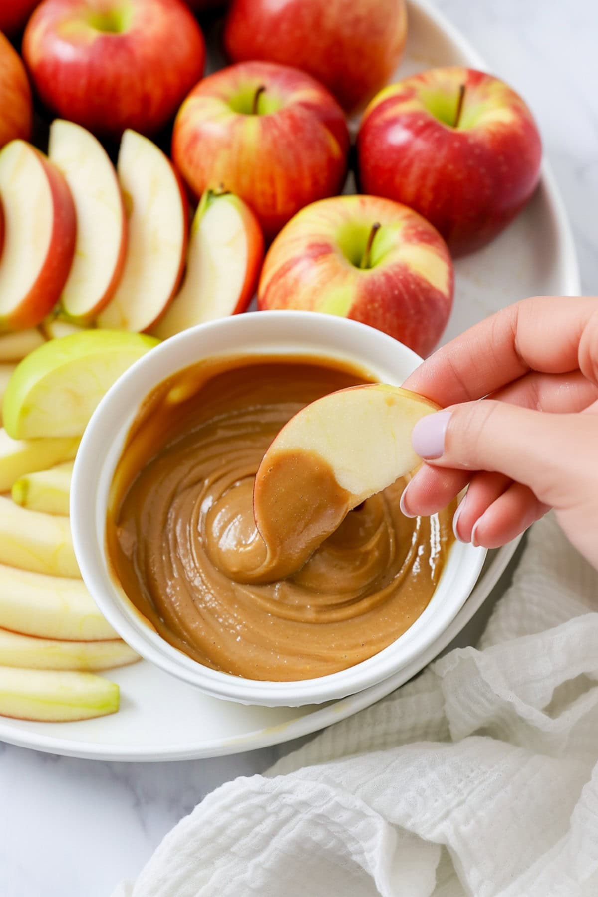 A hand dipping a slice of apple in a bowl of caramel dip on a white marble table