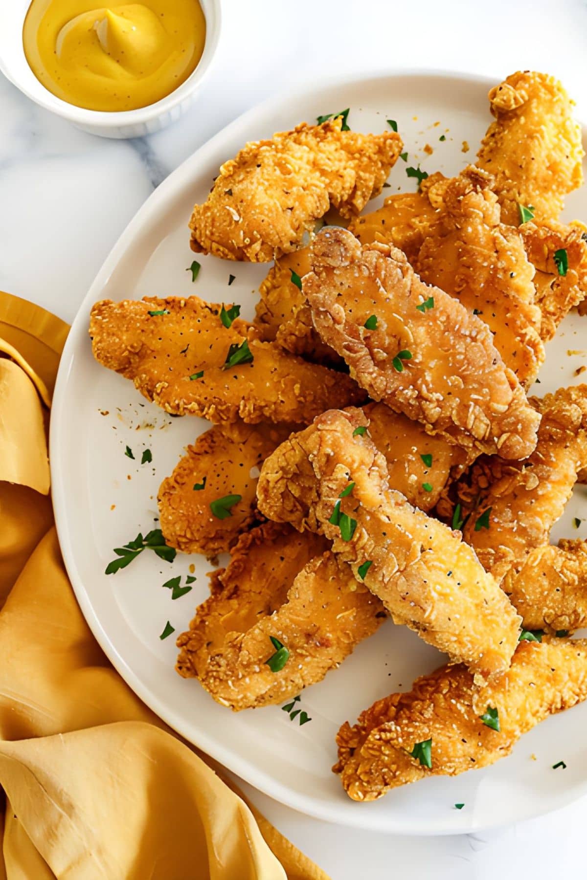 Close Up of Chicken Tenders with Parsley on a White Plate with a Ramekin of Honey Mustard and a Mustard Yellow Kitchen Towel