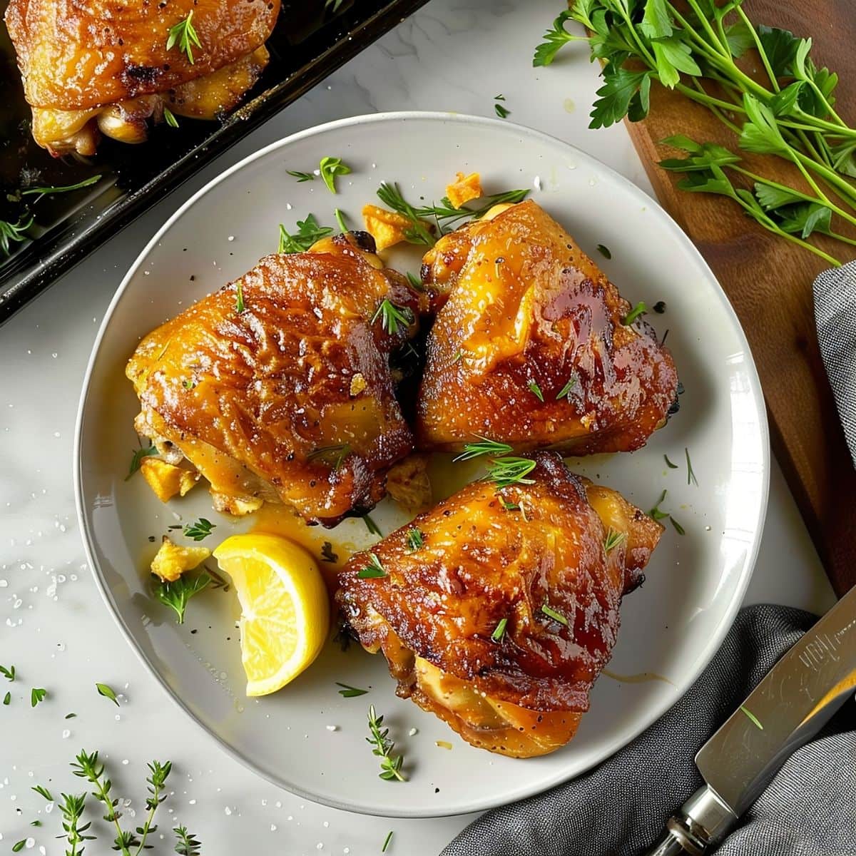 Three Baked Chicken Thighs on a Plate with Lemon on a White Marble Table with a Cutting Board and Tray of Chicken to the Sides