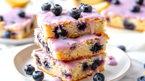 Slices of lemon blueberry blondies in a stacked in a saucer drizzled with light purple glaze.