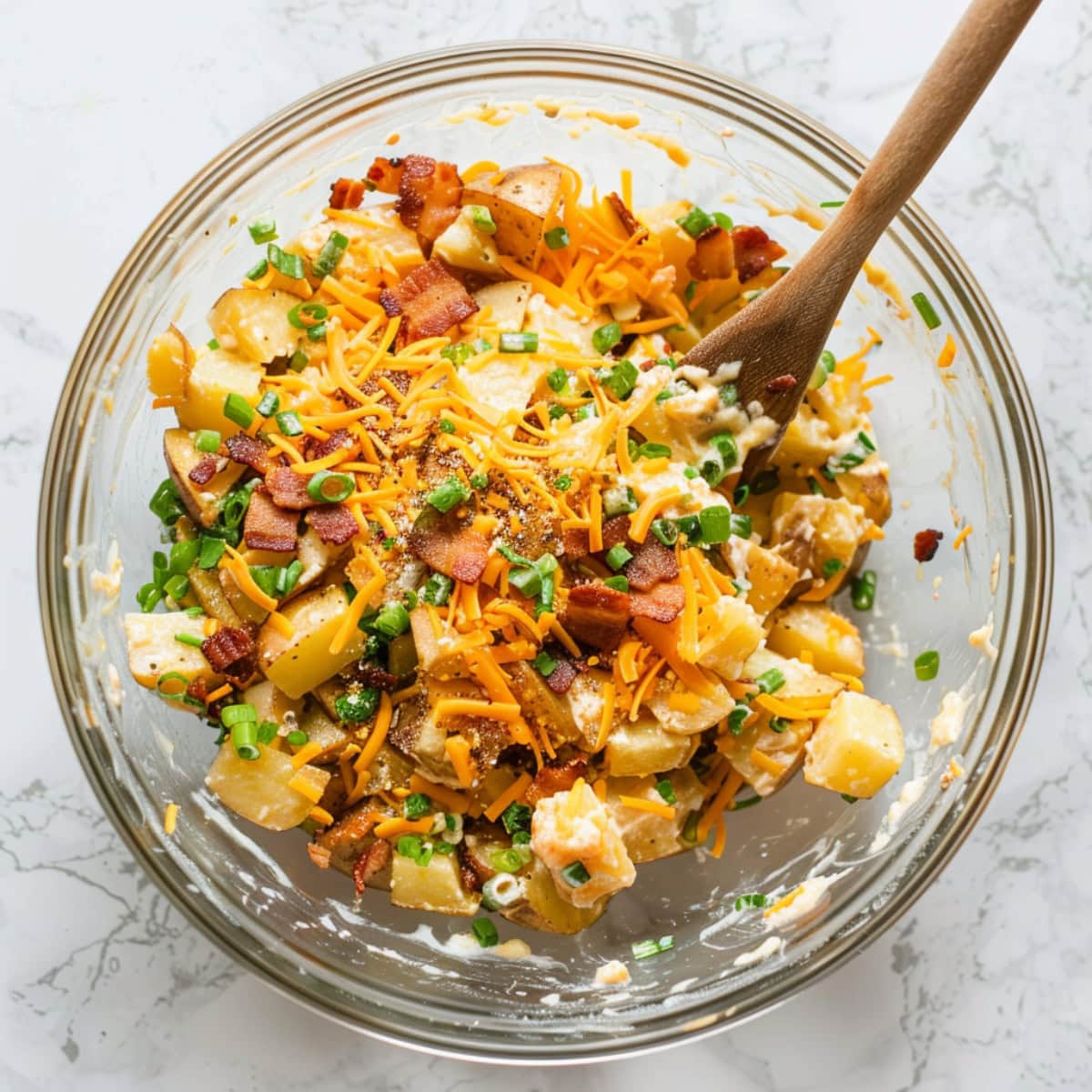 Peeled diced potatoes, grated cheddar cheese, crumbled bacon, sliced green onions, and garlic salt, mayonnaise mixed in a large glass bowl.