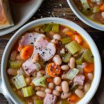 Two Bowls of Ham and Bean Soup with Meaty Chunks of Ham, Celery, Carrots, White Beans, and Onions on a Wooden Table with a Plate with Crusty Bread