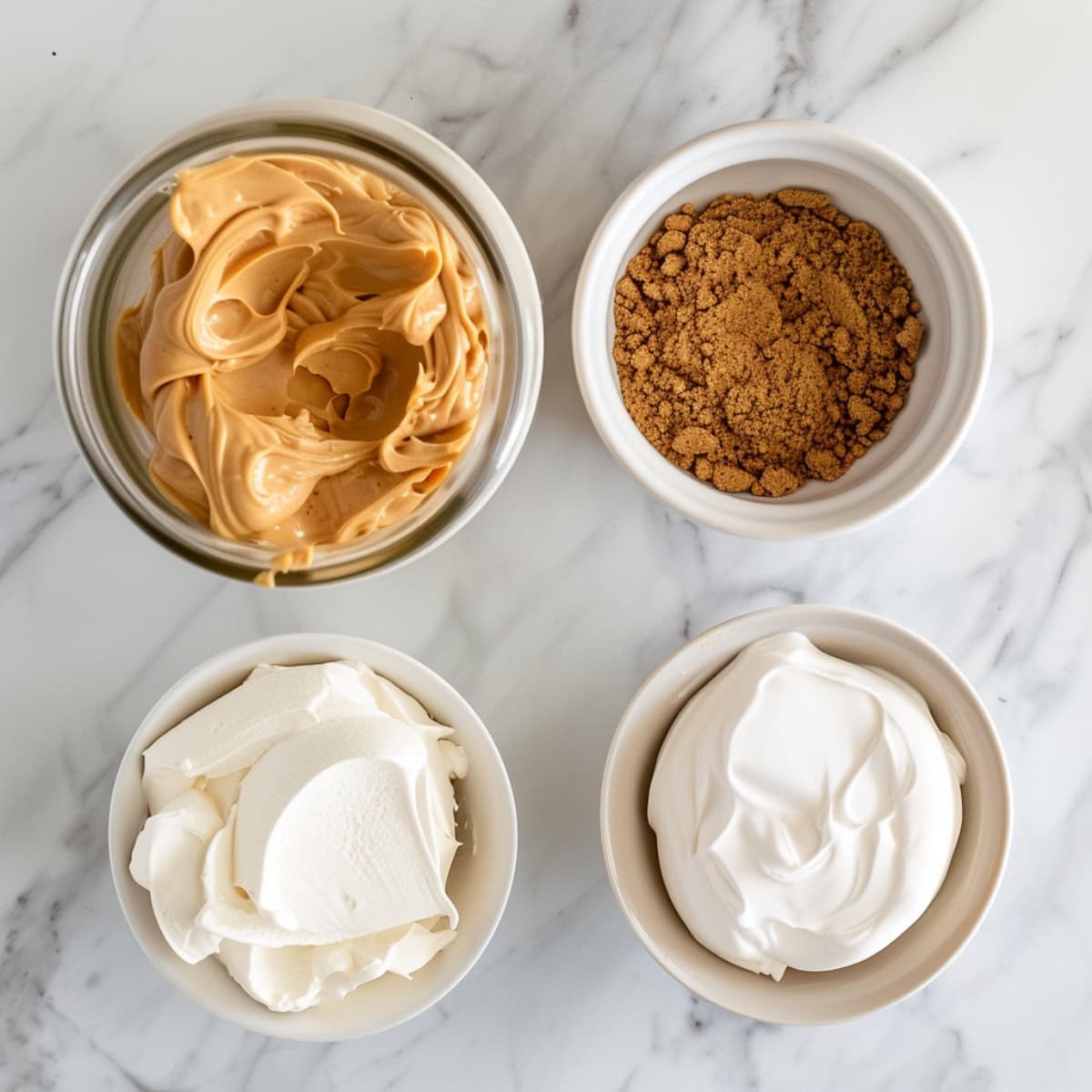 Peanut butter, brown sugar, whipped cream and heavy cream in bowls on a white marble table.