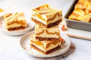 Square slices of pumpkin cheesecake bars stack in layer on a white plate.