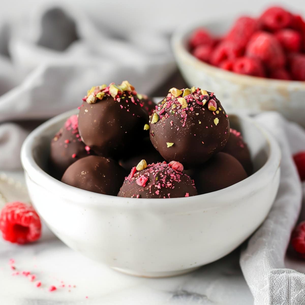 A set of beautifully arranged raspberry truffles in a white bowl topped with crushed pistachio nuts and freeze-dried raspberries