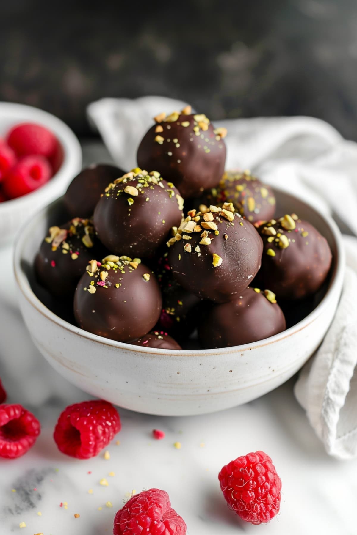 Chocolate raspberry truffles in a white bowl on a marble countertop with fresh raspberries on the side