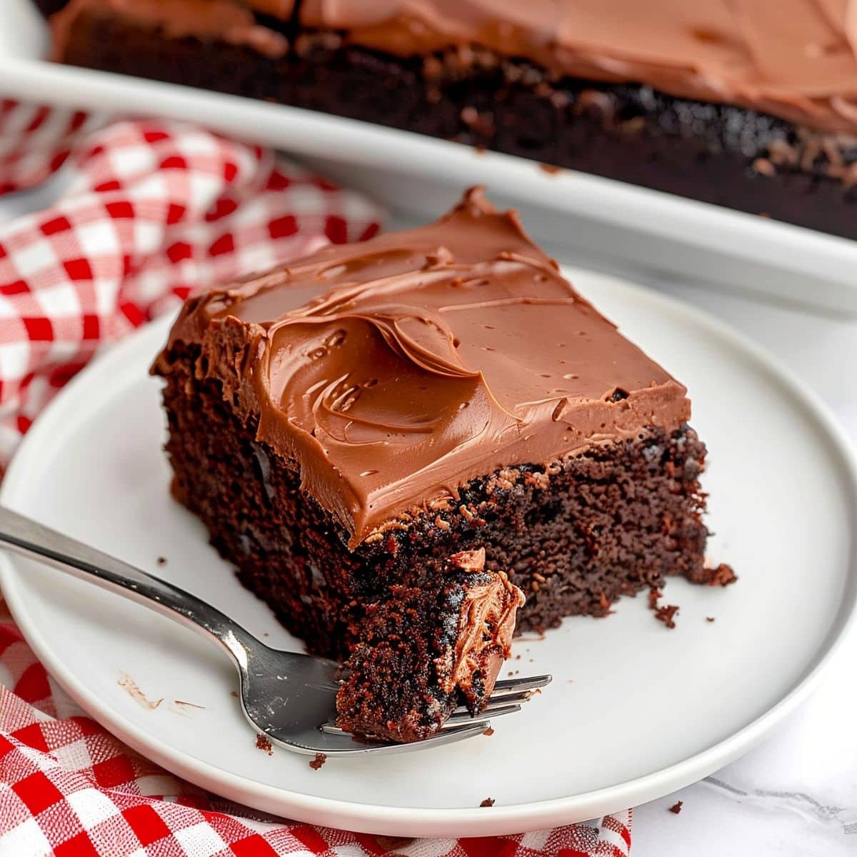 Slice of Chocolate Coca Cola Cake with Chocolate Frosting on a White Plate with a Fork, a Red Gingham Kitchen Towel, and the Rest of the Cake in the Tray Behind It