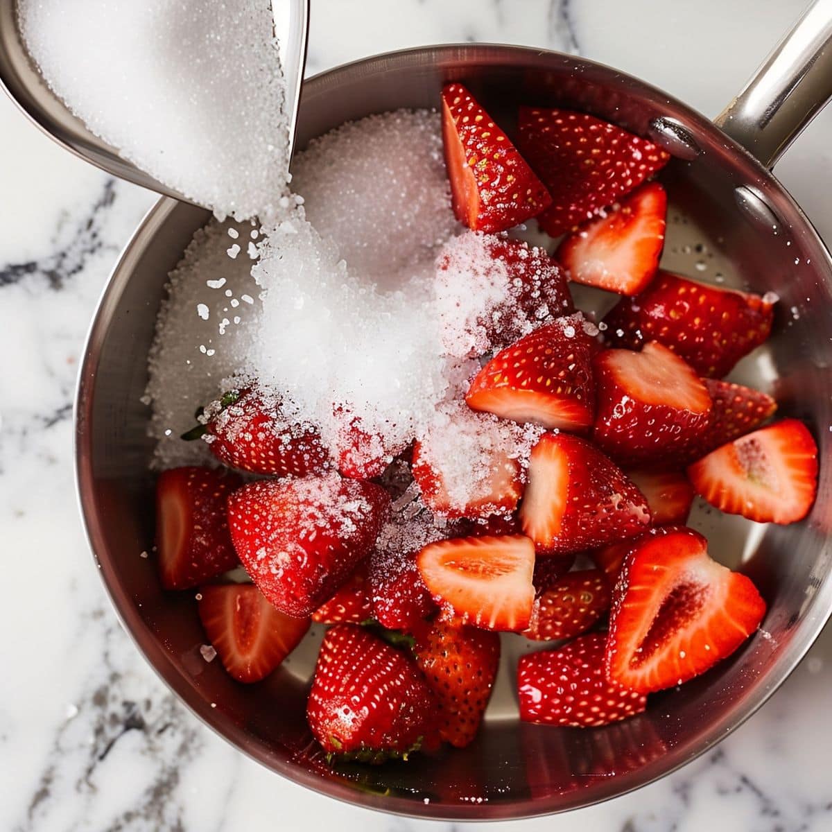 Top View of Saucepan with Halved Strawberries and Granulated Sugar on a White Marble Table