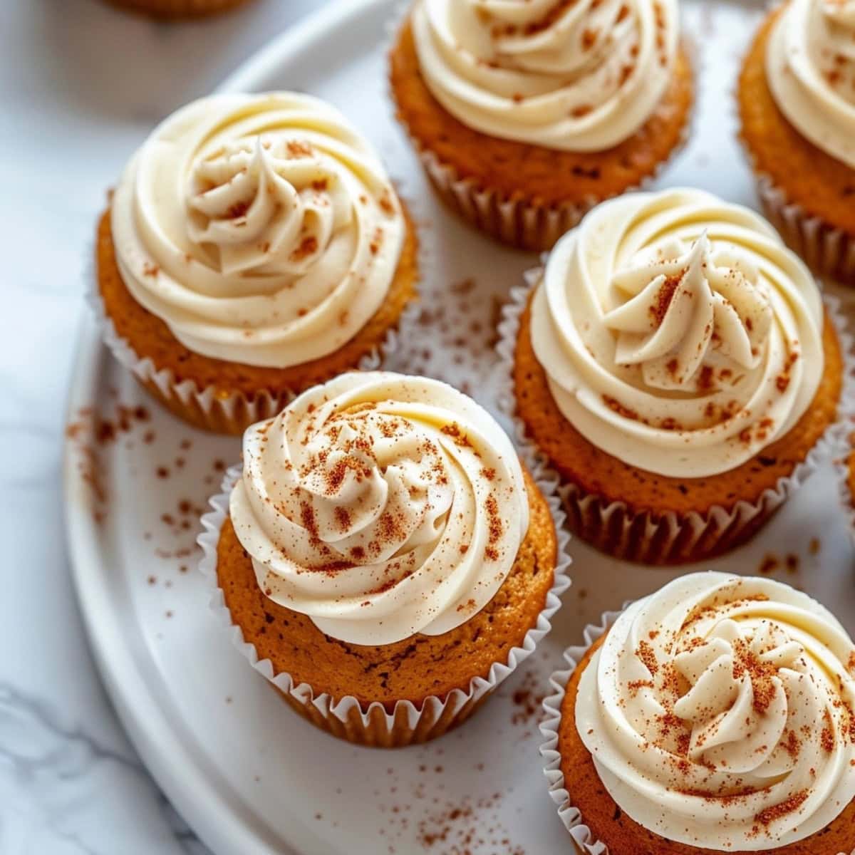 Pumpkin cupcakes with cream cheese frosting arranged on a white plate, sprinkled with pumpkin spice.