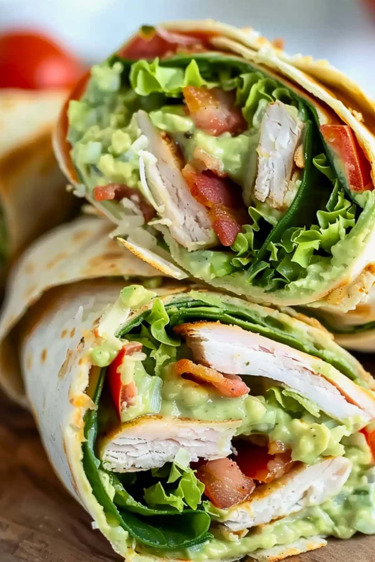 Sliced in half tortilla wrap with mashed avocado, turkey, tomatoes, bacon and shredded lettuce filling.