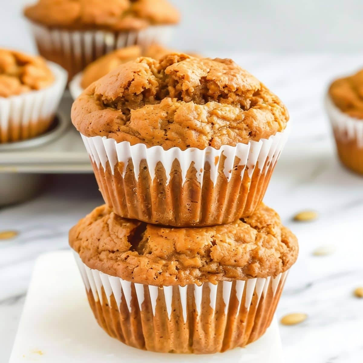Close Up of Two Two-Ingredient Pumpkin Muffins Stacked on a White Marble Table with a Tray of Muffins in the Background