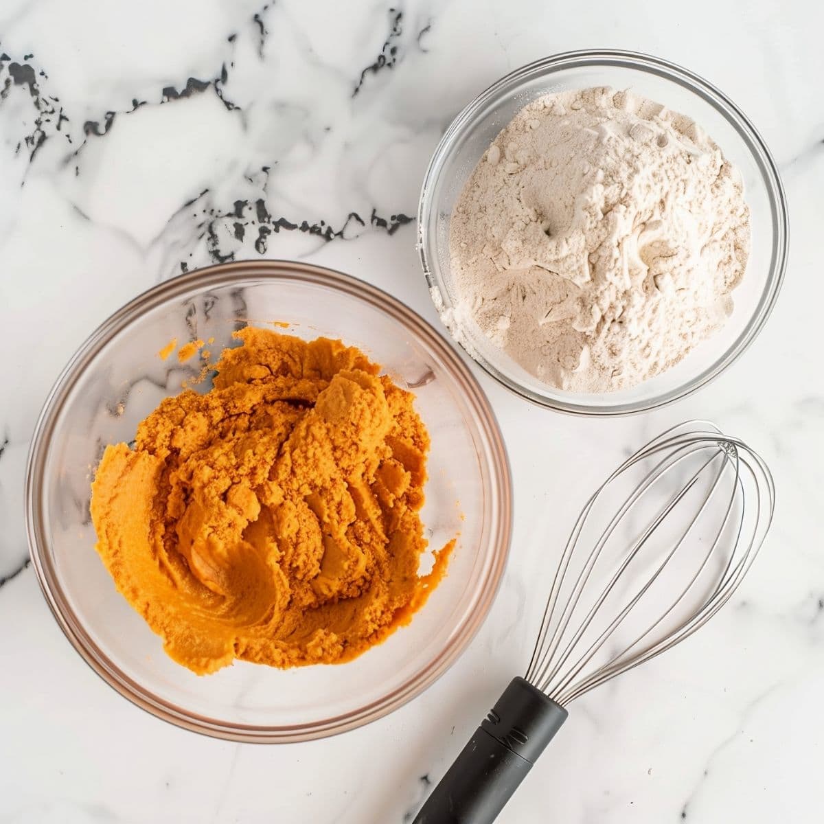 Pumpkin Puree in a Glass Bowl and a Spice Cake Mix in a Separate Glass Bowl on a White Marble Table with a Wire Whisk