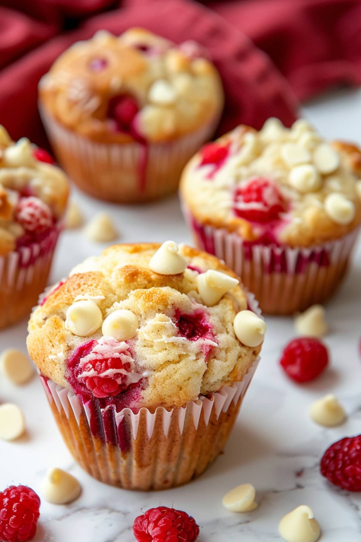 Raspberry and white chocolate  muffins, with scattered raspberries and mini white chocolate chips on a white marble surface.