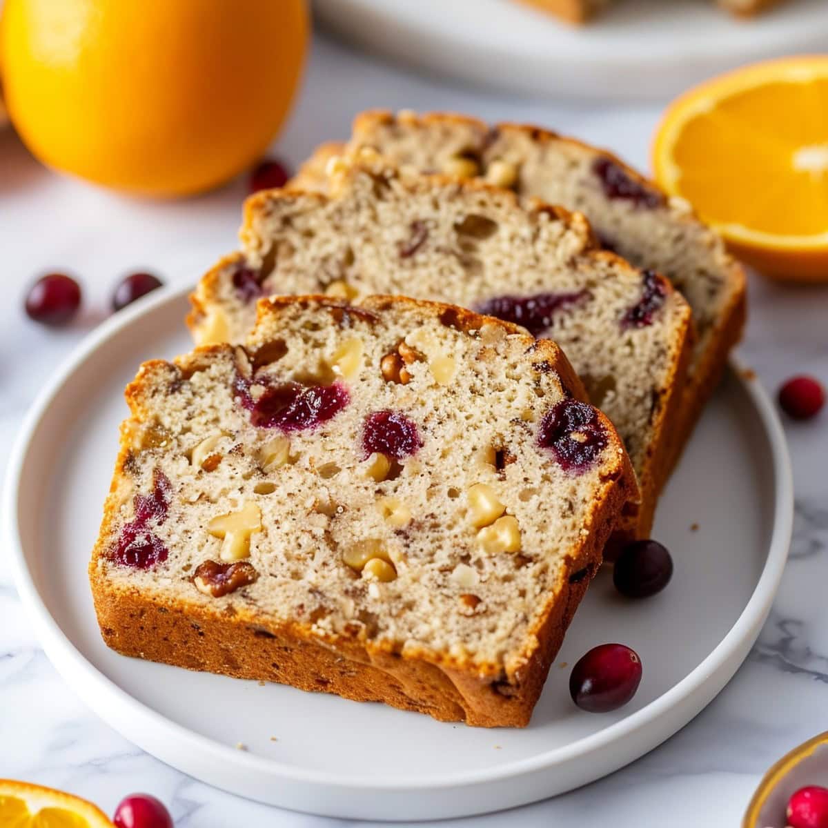 Three Slices of Cranberry Orange Bread on a White Plate with Cranberries and Oranges in the Background 