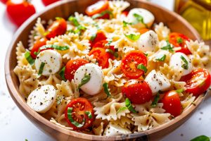 Caprese salad pasta in a wooden bowl with mozarella cheese balls, cooked farfalle pasta, cherry tomatoes in olive oil dressing.